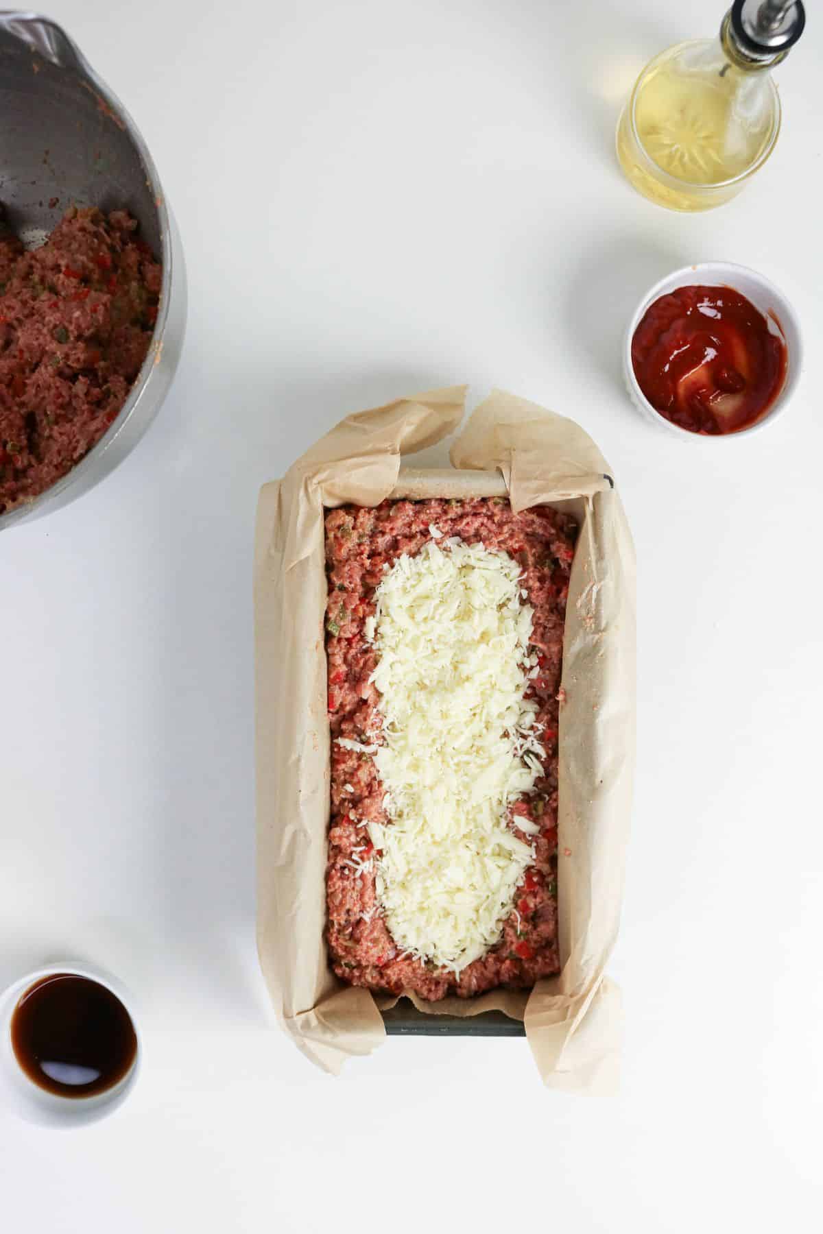 Uncooked meatloaf topped with shredded cheese in a parchment-lined loaf pan, surrounded by ingredients.