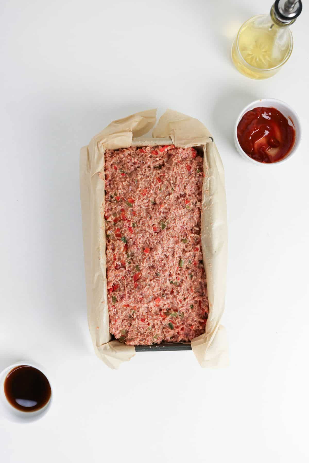 Unbaked meatloaf in a parchment-lined loaf pan with small bowls of sauce nearby.