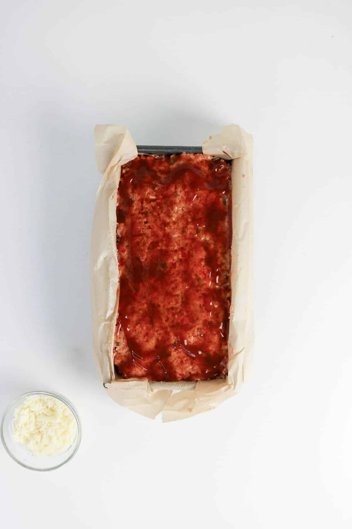 A loaf of meat in a parchment-lined pan topped with glaze, next to a small bowl of shredded cheese.