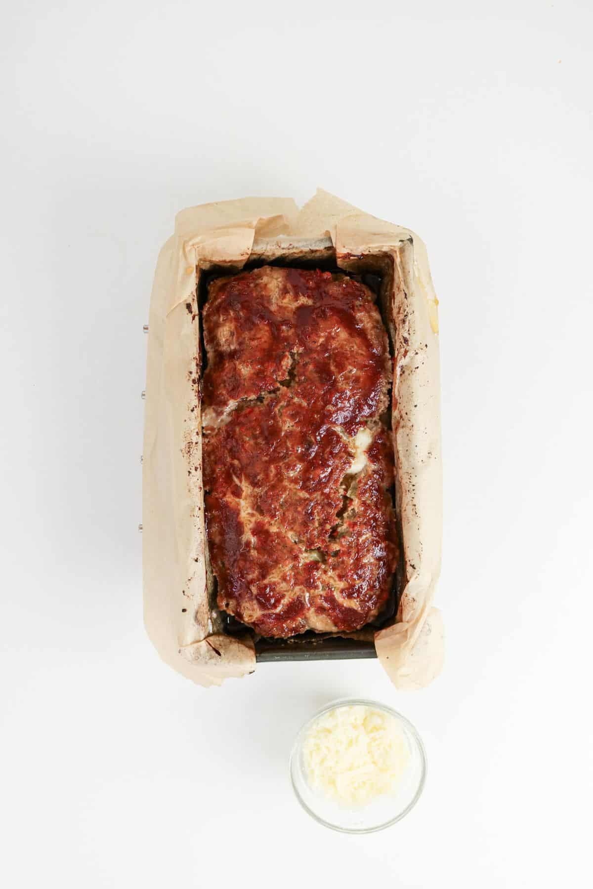 Glazed meatloaf in a parchment-lined loaf pan, with a small bowl of shredded cheese nearby.