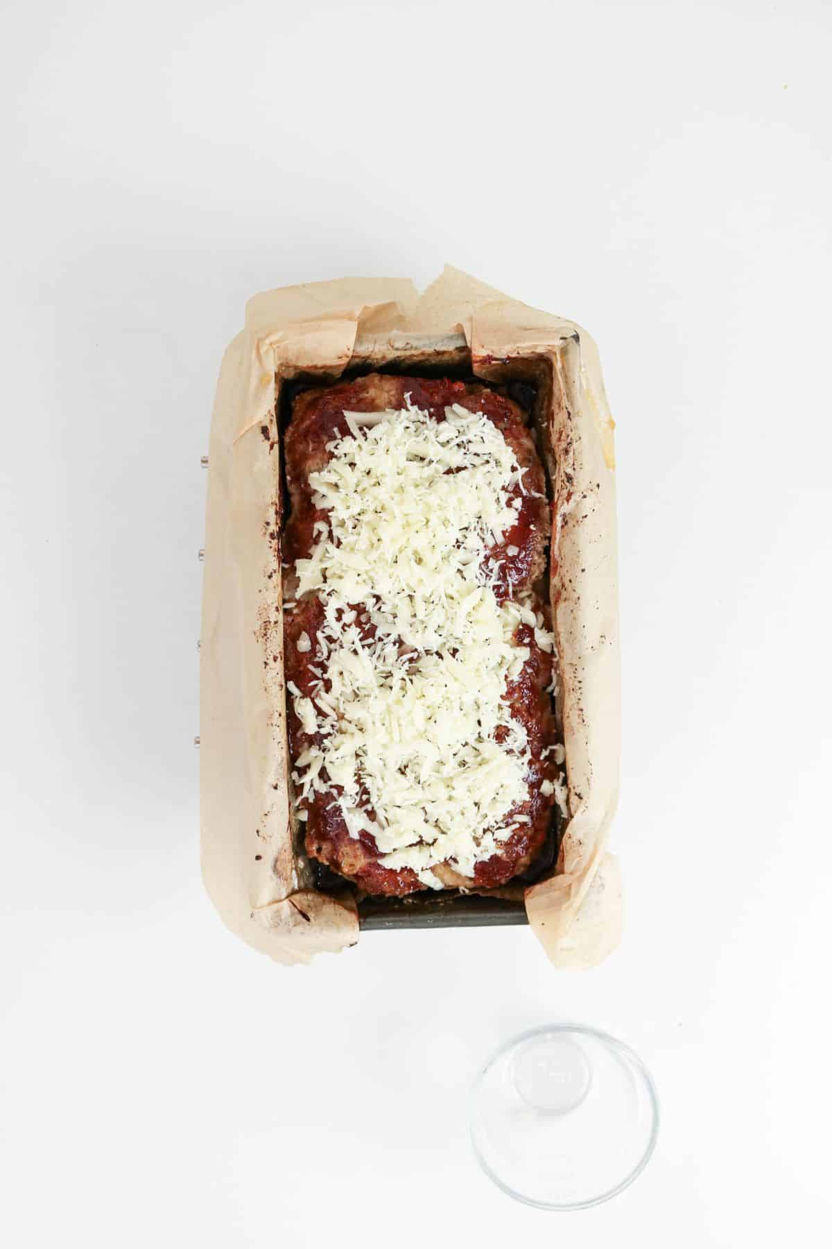 Meatloaf topped with ketchup and shredded cheese in a parchment-lined loaf pan.