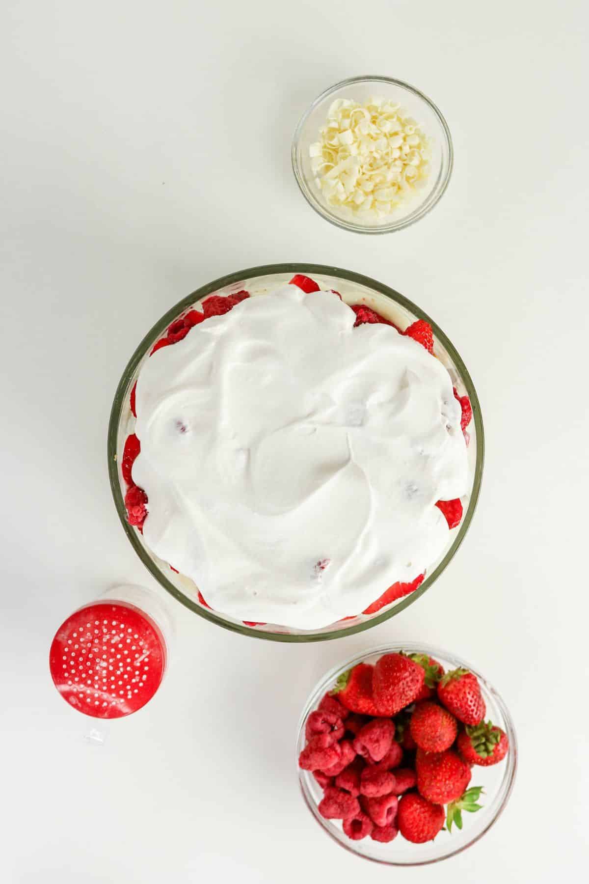 The final layer of whipped topping, with berries, white chocolate curls, and a powdered sugar shaker ready for garnishing.